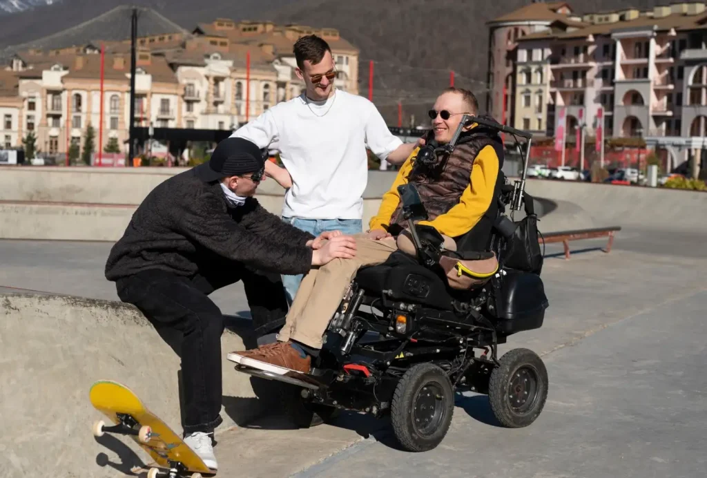 Three friends hanging out at a skatepark, one in a wheelchair, enjoying a sunny day with urban buildings in the background.