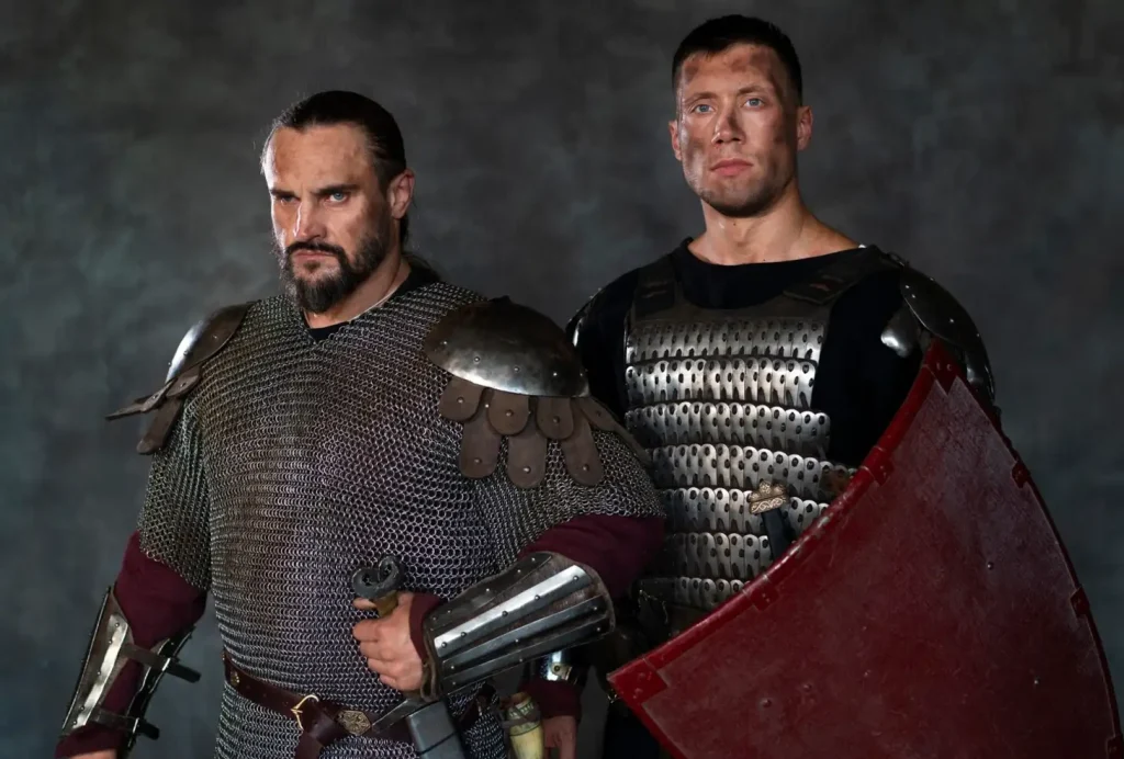 Two medieval warriors in chainmail and metal armor stand confidently, one holding a sword and the other a large shield.