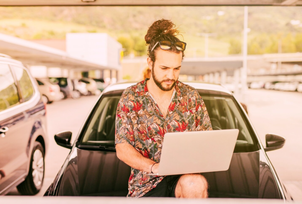 Man with bun using laptop on car hood in parking lot, wearing sunglasses and floral shirt.