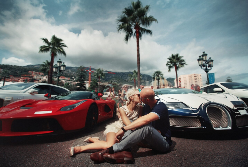 Couple kissing on a street with luxury cars, palm trees, and buildings in the background, under a cloudy sky.