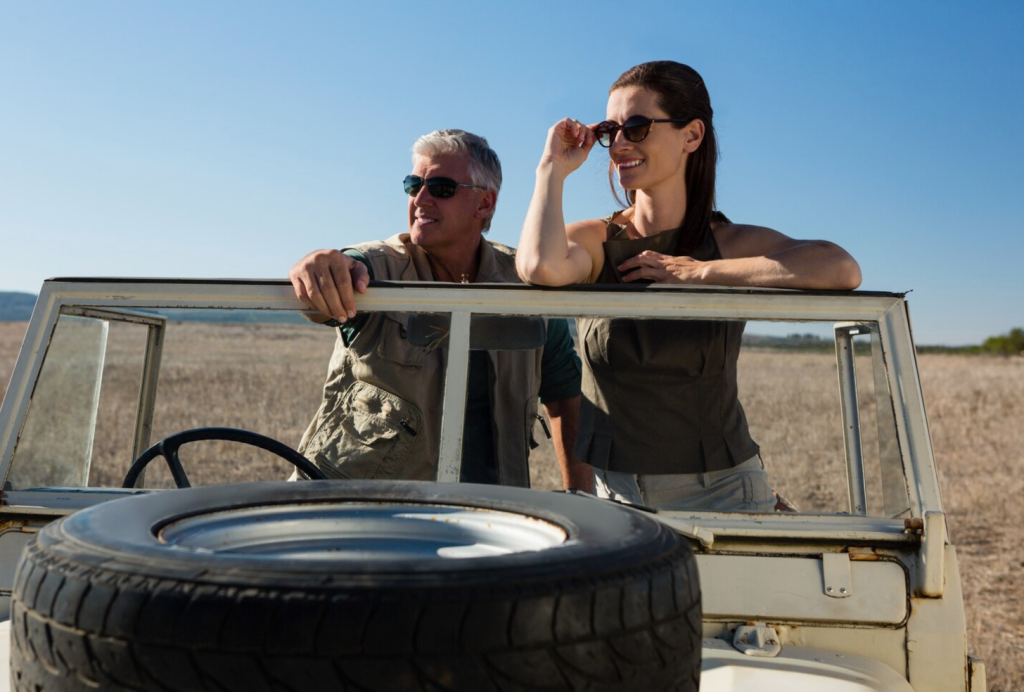 Two people in sunglasses standing by an open-top jeep in a dry field under a clear blue sky.