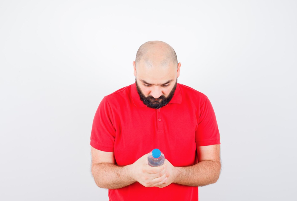 Man in a red shirt looking down at a water bottle in a bright, white background.