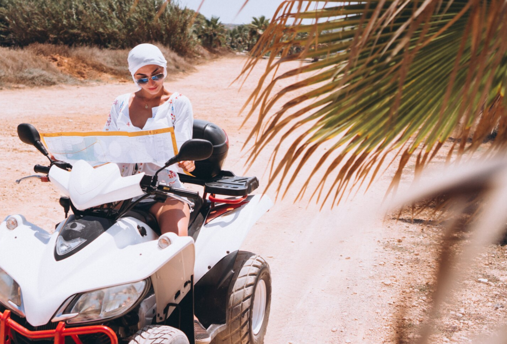 Person reading a map on a parked quad bike by a dirt road, surrounded by palm leaves and sunny landscape.