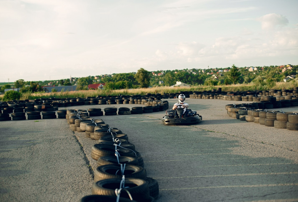 Person driving go-kart on outdoor track, surrounded by stacks of tires, with a scenic background of trees and houses.