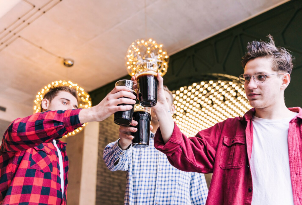 Three people clinking glasses of dark beer in a festive setting with decorative lights.