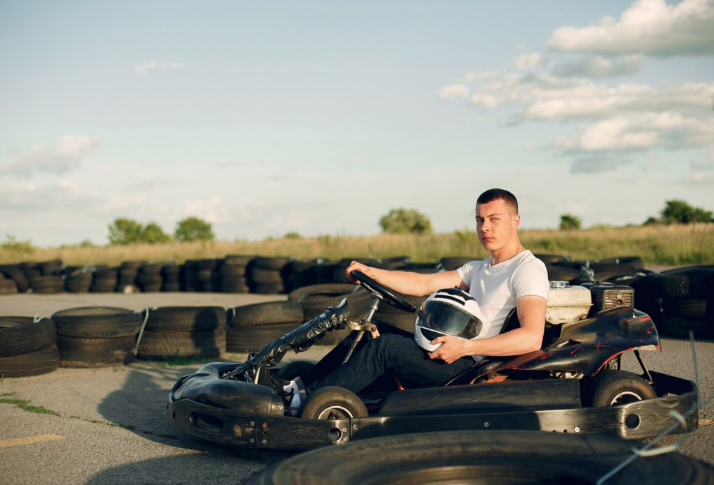 Man in a white shirt holding a helmet, sitting in a go-kart on a track with stacked tires under a blue sky.
