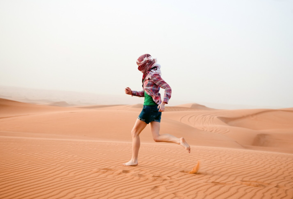 Person running barefoot on sand dunes under a clear sky, wearing a headscarf, plaid shirt, and shorts.