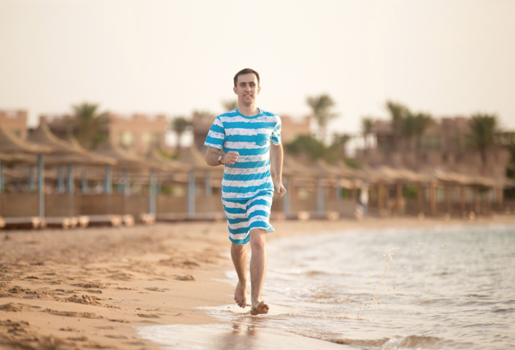 Man in striped shirt running on sandy beach by the water, with beach huts in the background.