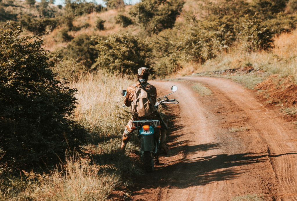 A person rides a motorcycle on a dirt path through hilly, green countryside, surrounded by trees and grass.