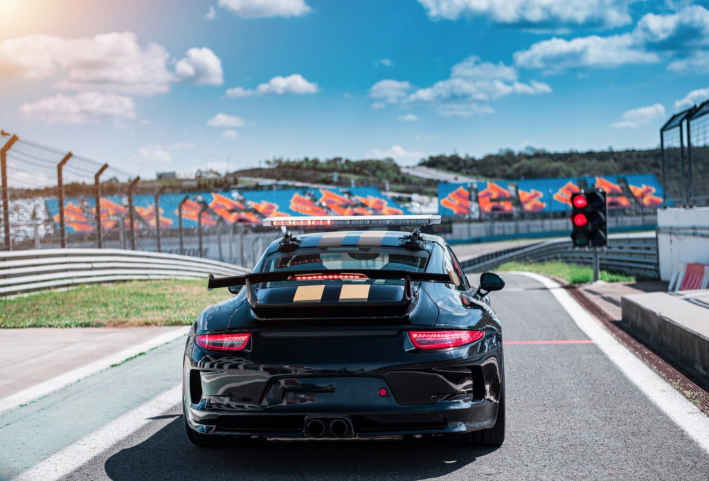 Black race car on a track with red tail lights, blue sky, and colorful banners in the background.