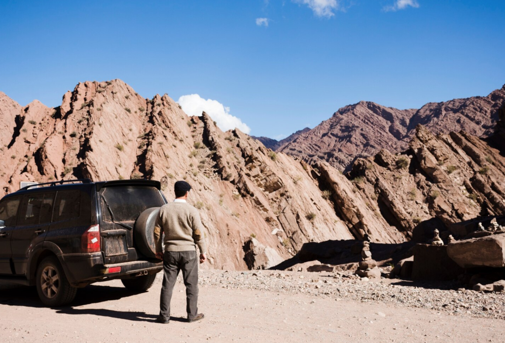 Man stands by SUV on rocky mountain road, gazing at rugged red peaks under a clear blue sky.