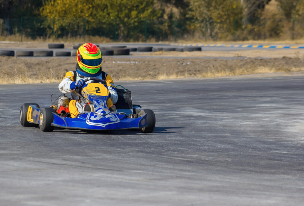 Person in a helmet driving a blue go-kart on a racetrack, surrounded by tires and trees in the background.