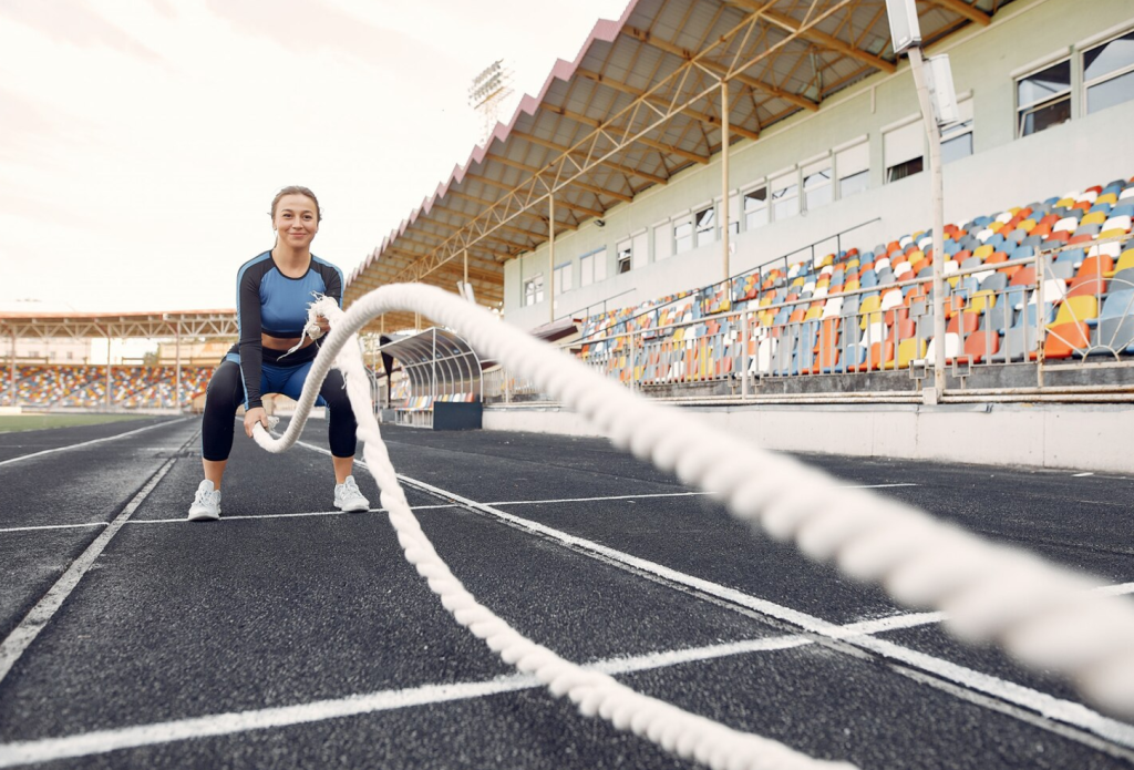 Woman exercising with battle ropes on a track in front of colorful stadium seats.