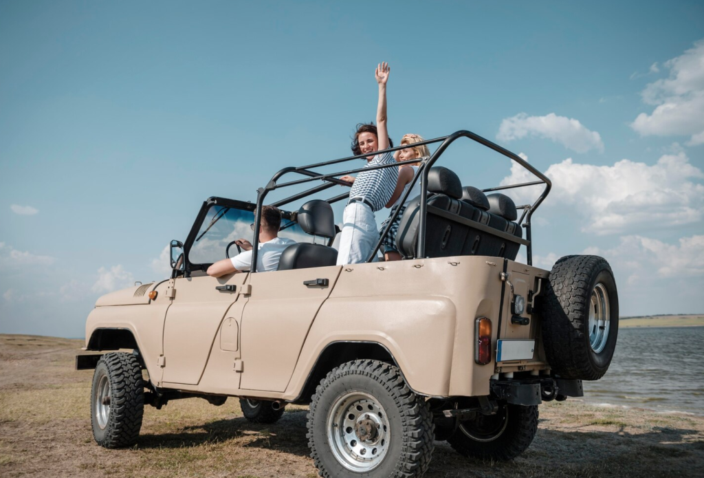 Group enjoying a sunny day in an open-top jeep by a lake, with one person standing and waving.