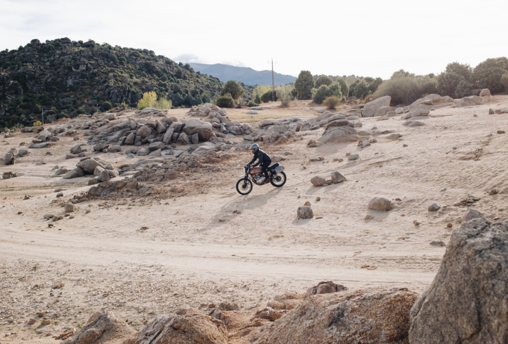 A person rides a motorcycle on a rocky desert trail, surrounded by boulders and sparse vegetation.