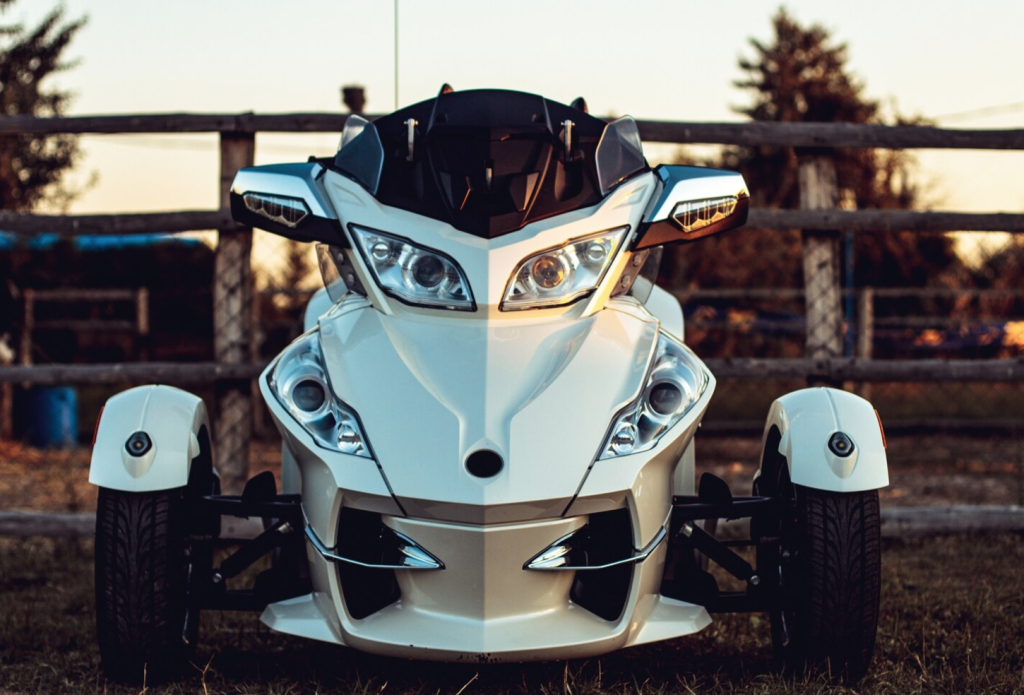 Front view of a white three-wheeled motorcycle with dual headlights, parked on grass against a wooden fence, under evening sky.