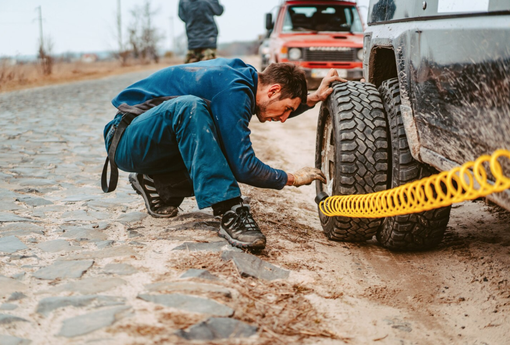 Man kneeling on a cobblestone road, inflating a car tire with a yellow air hose. Red vehicle in the background.