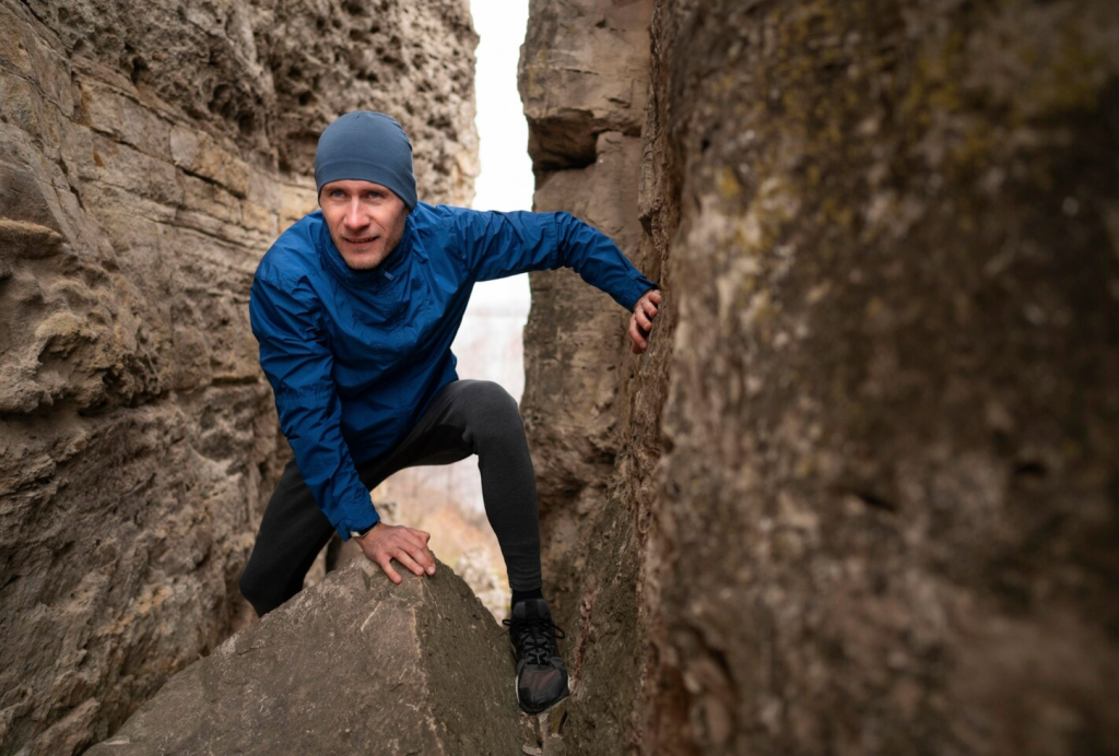 Man in blue jacket climbing between large rock formations outdoors.