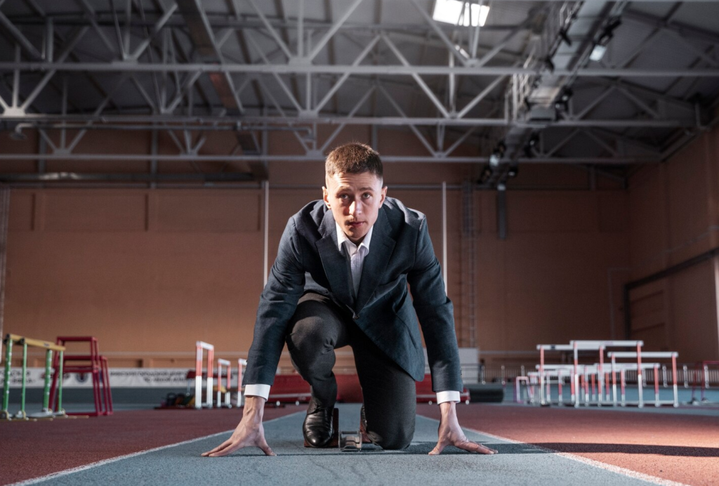 Man in a suit crouching at a starting block on an indoor track, ready to sprint.