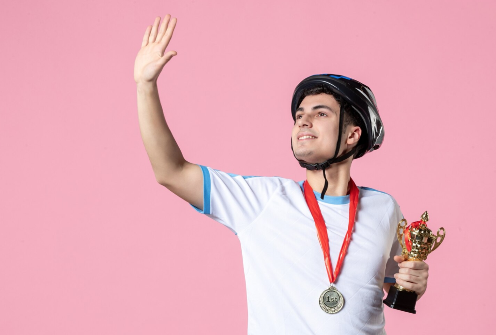 Smiling person in a helmet holds a trophy and medal, raising their hand against a pink background.