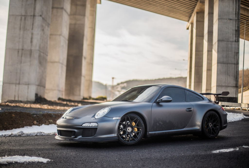 Matte gray sports car parked under a bridge on a sunny day.