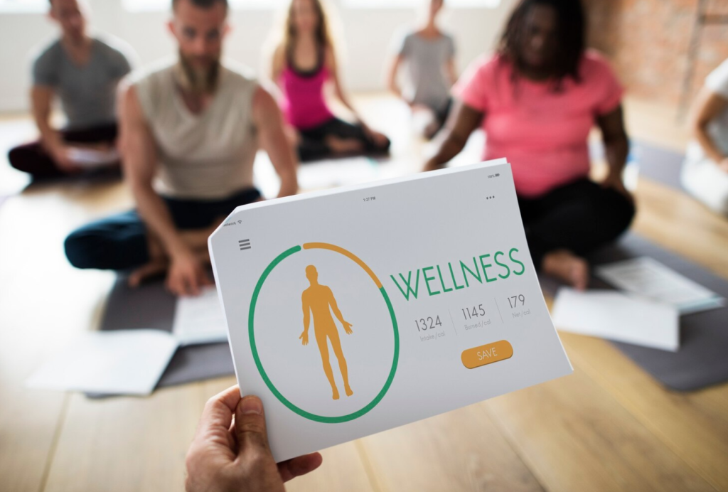 Person holds a wellness app displaying calories over a yoga class with people sitting on mats in the background.