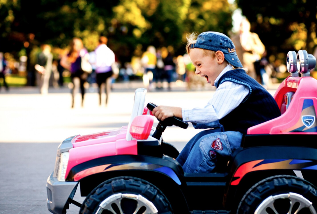Child joyfully drives a pink toy car in a park, wearing a blue cap and jacket, with blurred people in the background.
