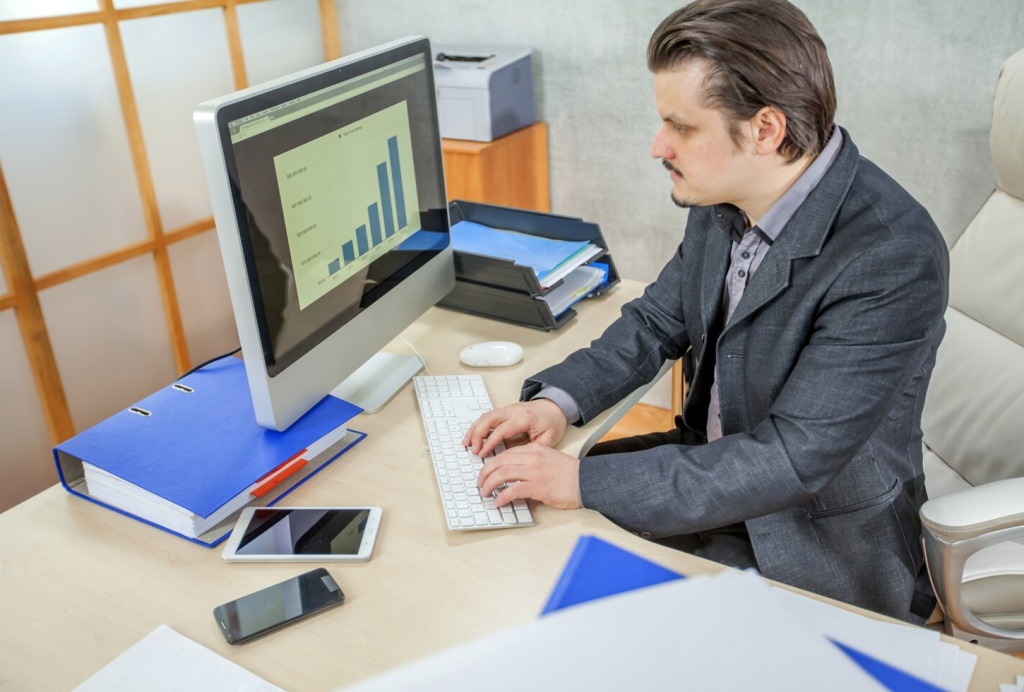 Man in a suit working at a desk with a computer displaying a bar graph, surrounded by office supplies.