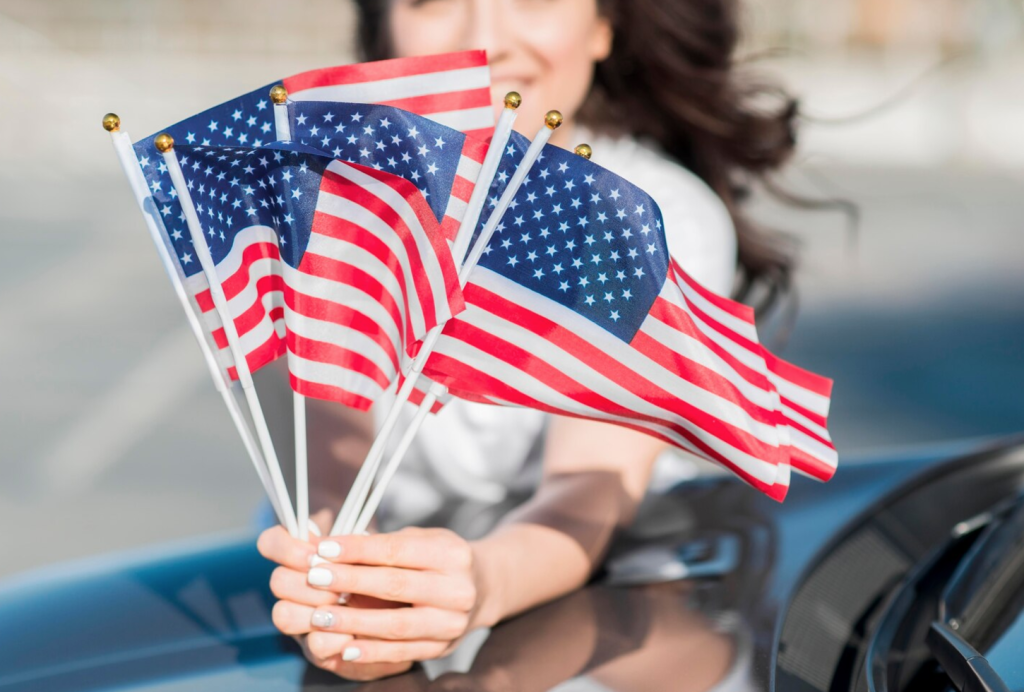 A person holds several small American flags, with a blurred background and partial view of a car hood.