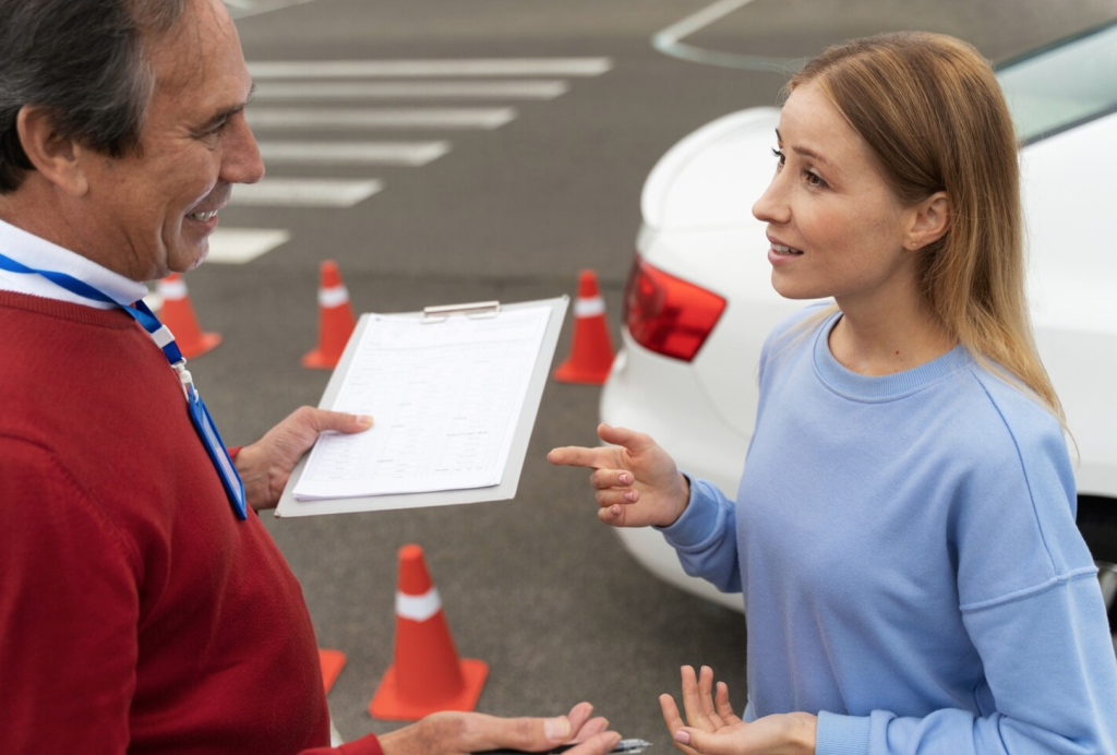 Instructor and woman in conversation near car, with clipboard and traffic cones in the background.