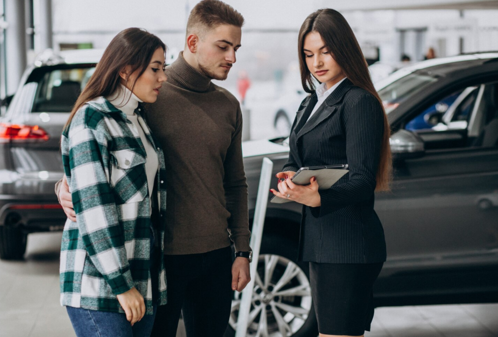A couple consults with a car dealer inside a showroom, standing in front of a gray vehicle.