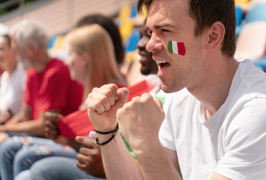 Enthusiastic soccer fan with Italian flag face paint cheers in a crowd, wearing a white shirt.