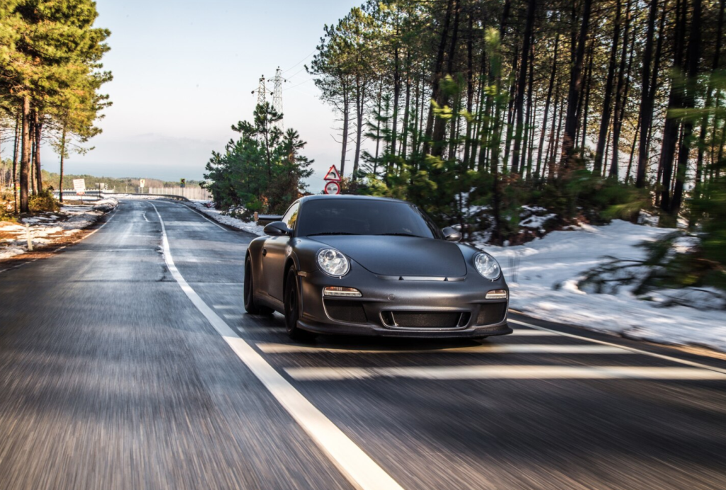 A sleek black sports car driving down a snowy forest road under a clear sky.
