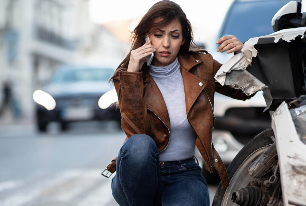 A worried woman in a brown jacket talks on the phone beside a damaged car on a busy street.