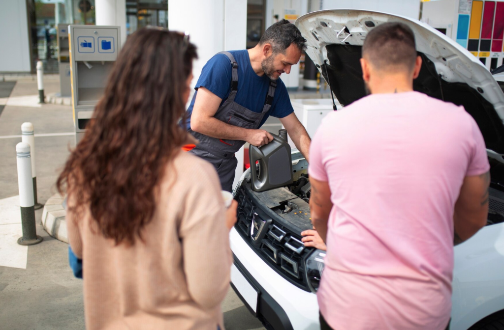 Mechanic pouring oil into a car engine while two people watch attentively at a service station.