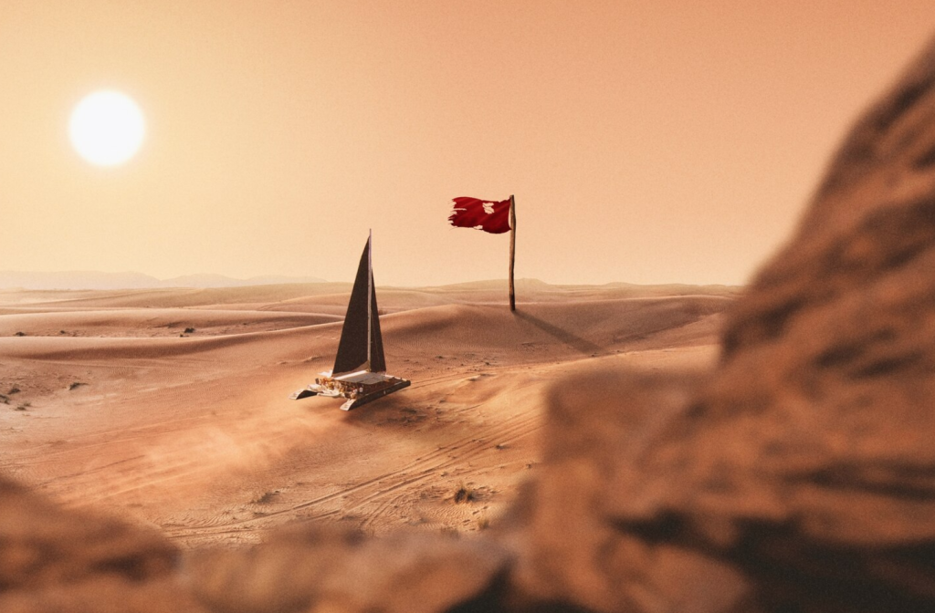 A sailboat glides across a desert landscape under a bright sun, with a tattered red flag on a pole in the background.