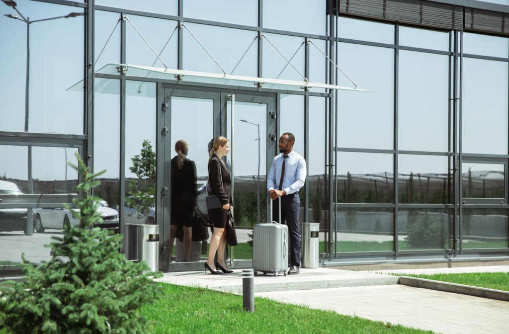 Two professionals with a suitcase converse outside a modern glass building on a sunny day.