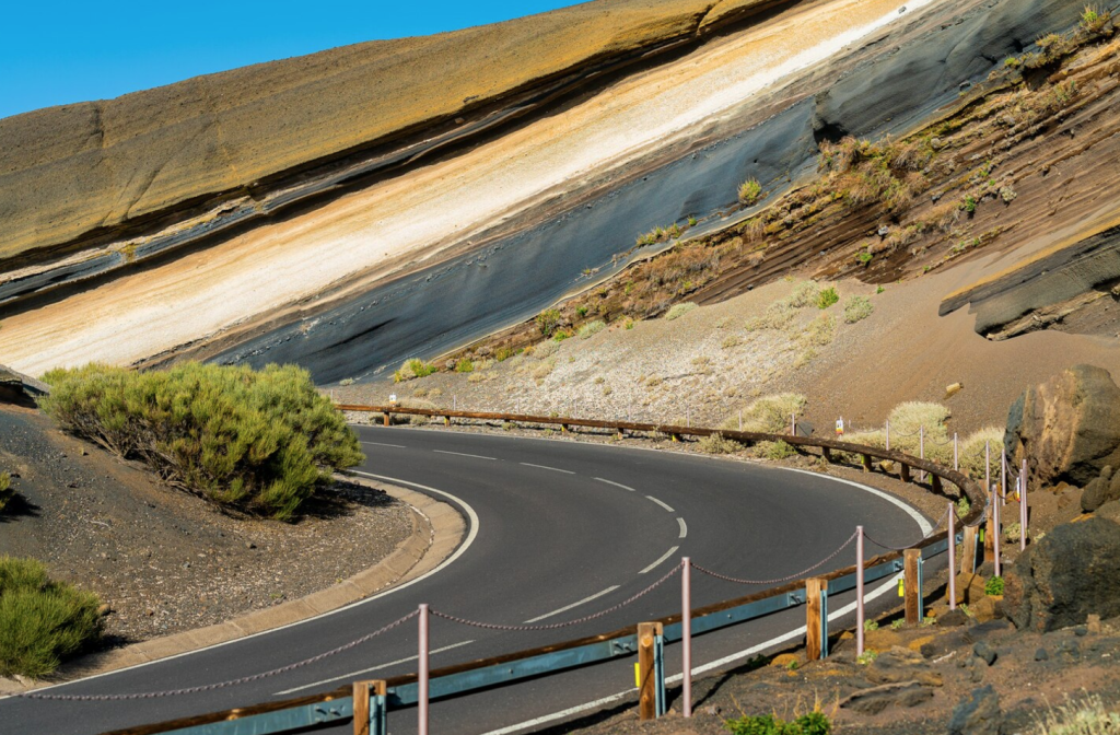 Scenic winding road through layered volcanic rock formations with desert vegetation under a clear blue sky.