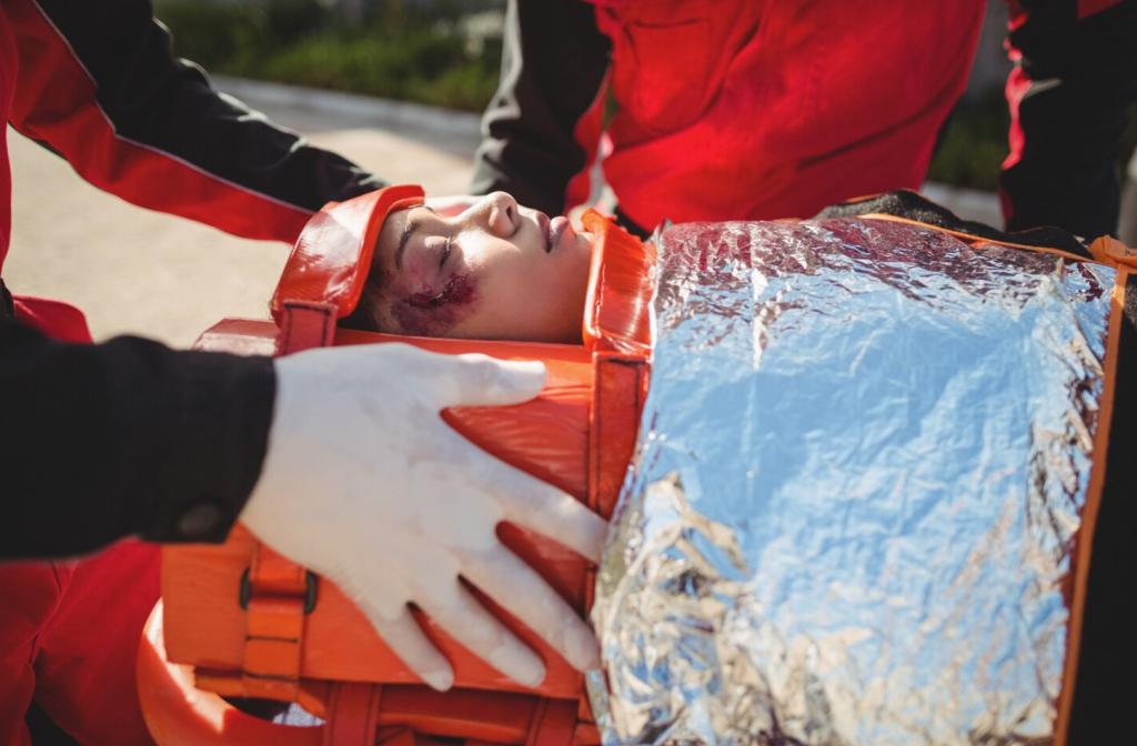Injured person on stretcher with head brace and foil blanket, attended by emergency responders in red uniforms.