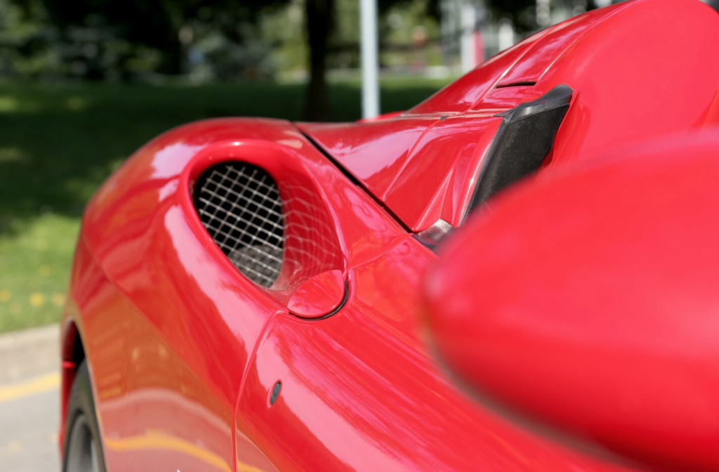 Close-up of a red sports car's side air intake with a mesh grille, reflecting sunlight in a green outdoor setting.